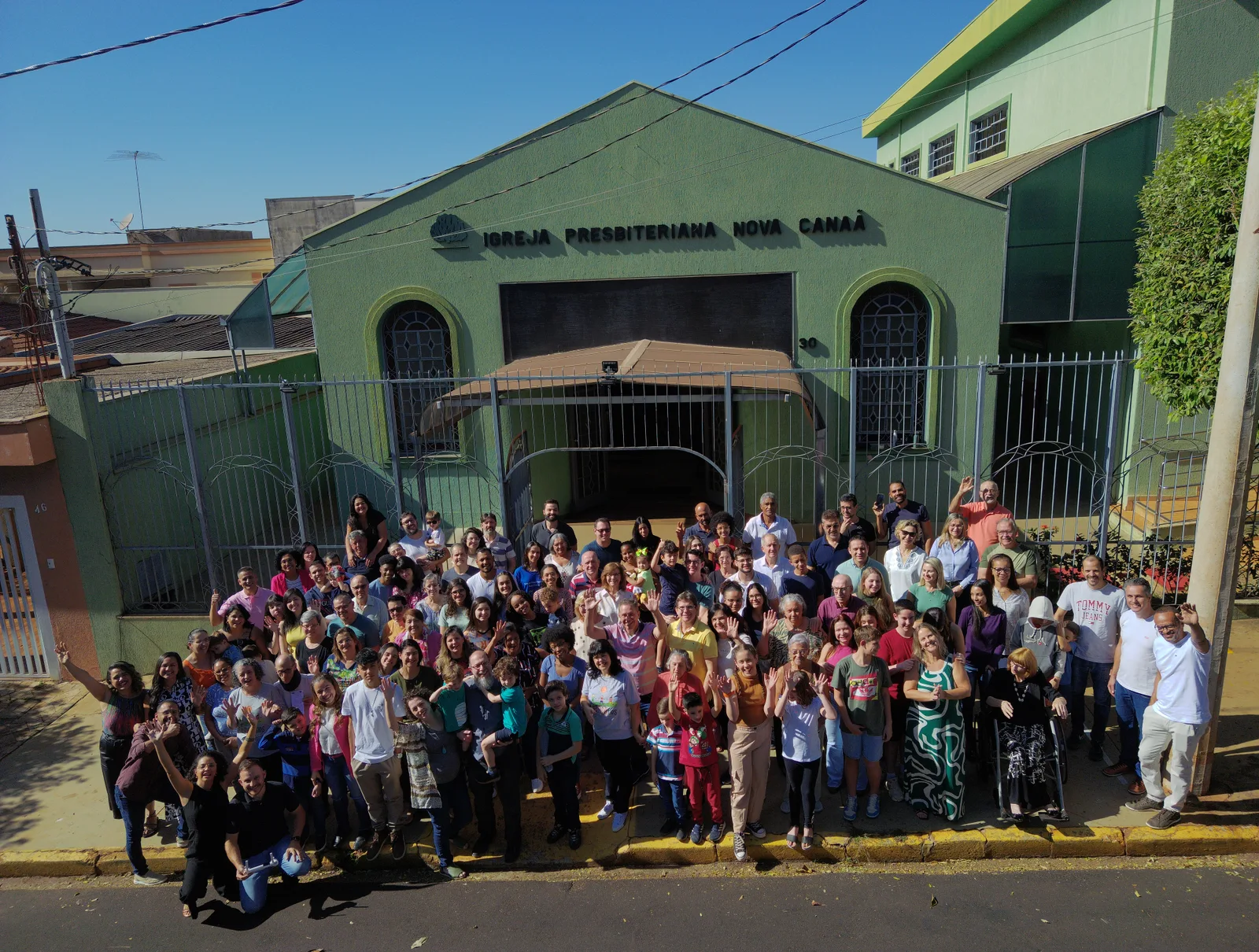 Membros da Nova Canaã reunidos em frente à fachada da igreja, acenando e sorrindo